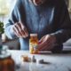 Eine ältere Person steht in einer Küche und hält eine durchsichtige Medikamentenflasche mit verschiedenen Tabletten in den Händen. Auf der Arbeitsfläche und im Hintergrund sind weitere Medikamentenbehälter zu sehen.