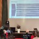 In einem großen Vortragssaal steht eine Frau im Business-Outfit an einem Rednerpult mit der Aufschrift „Universität Freiburg“ und spricht vor einem sitzenden Publikum. Auf einer großen Leinwand hinter ihr wird eine Präsentation gezeigt, die sich mit dem „DFG Kodex“ zur wissenschaftlichen Integrität befasst; sichtbar sind mehrere Leitlinien zu Qualitätssicherung, Verantwortung, Forschungsdesign und Dokumentation. Links neben dem Pult steht ein Aufsteller mit dem Logo von Cochrane Deutschland, rechts auf der Bühne ein großer weißer Blumenstrauß in einer Vase.