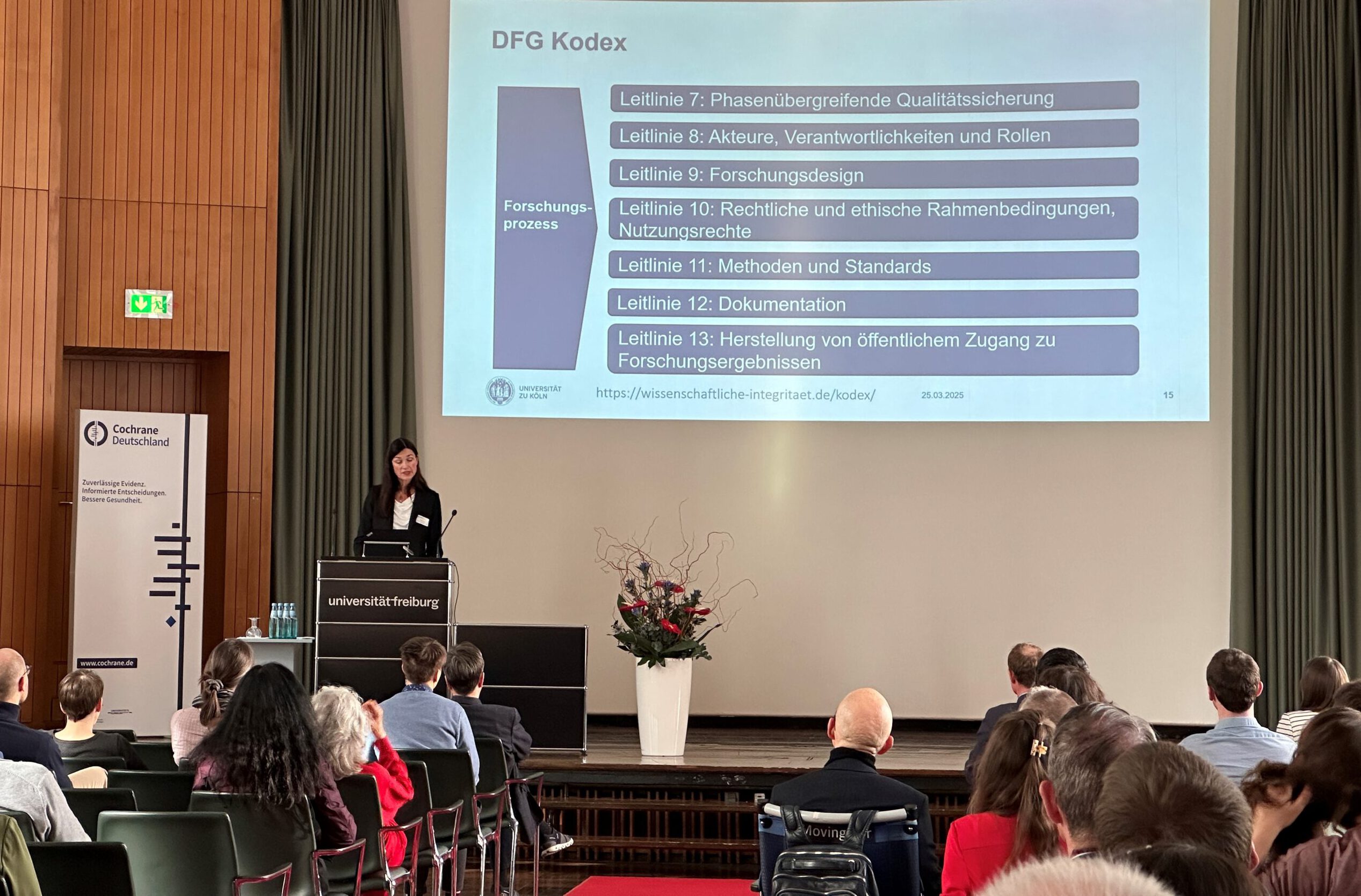 In einem großen Vortragssaal steht eine Frau im Business-Outfit an einem Rednerpult mit der Aufschrift „Universität Freiburg“ und spricht vor einem sitzenden Publikum. Auf einer großen Leinwand hinter ihr wird eine Präsentation gezeigt, die sich mit dem „DFG Kodex“ zur wissenschaftlichen Integrität befasst; sichtbar sind mehrere Leitlinien zu Qualitätssicherung, Verantwortung, Forschungsdesign und Dokumentation. Links neben dem Pult steht ein Aufsteller mit dem Logo von Cochrane Deutschland, rechts auf der Bühne ein großer Blumenstrauß in einer weißen Vase.